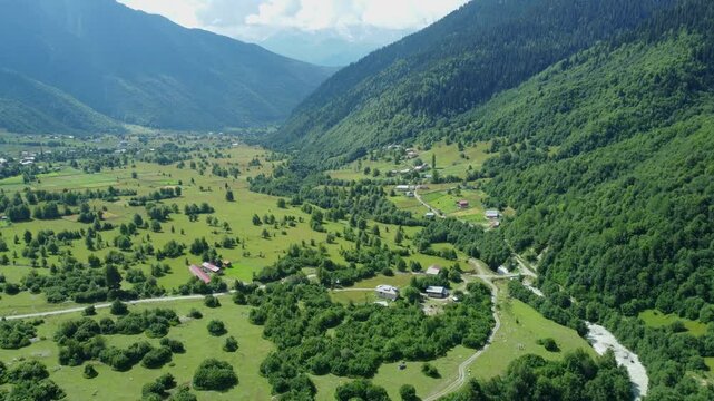 White clouds gently caress towering mountain peak, casting shadows on bright green valley below, where dirt road winds through picturesque landscape. Shooting from drone.