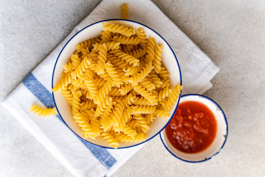 Overhead view of a bowl of Raw fusilli pasta and a bowl of tomato sauce on a table