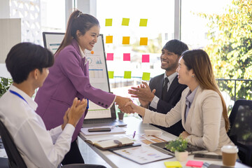 A group of people are gathered around a white board with a woman giving a presentation. Scene is positive and collaborative, as the group is clapping and smiling
