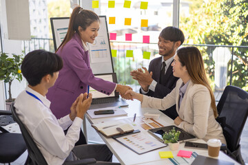 A group of people are gathered around a white board with a woman giving a presentation. Scene is positive and collaborative, as the group is clapping and smiling
