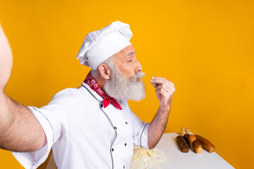 Cheerful senior chef wearing uniform presenting culinary skills with bread loaves and flour-filled countertop against yellow background