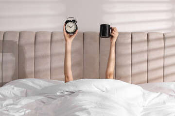Woman with alarm clock and cup in bed at home, closeup. Good morning
