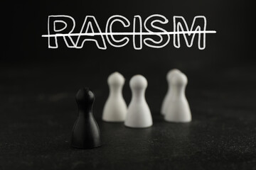 Stop racism. Black game piece standing apart from white ones on table. Strikethrough word