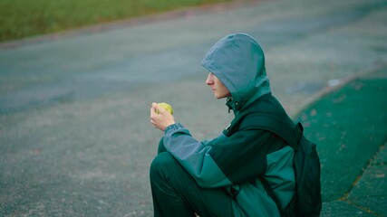 Young man sitting on ground eating green apple outdoors in residential area. Male teenager taking bite from apple while seated near street and greenery. Youth having snack on pavement in quiet