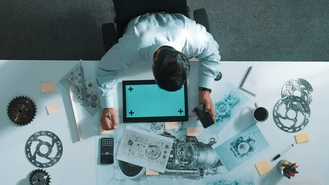 Top view of engineer looking at phone while working on tablet with green screen at meeting table with turbine engine or jet engine sketch design and various metal gear scatter around. Alimentation.