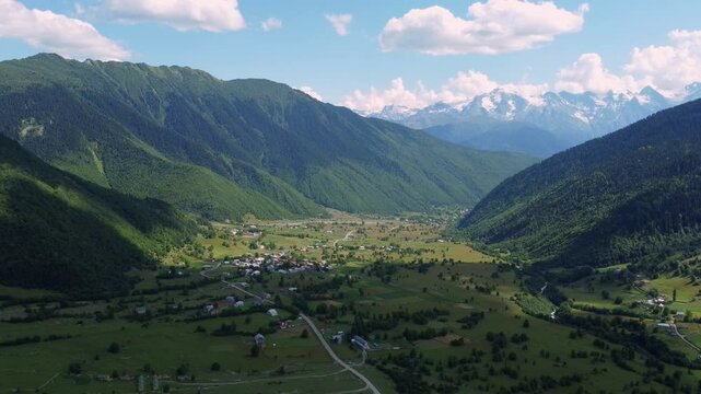 White clouds gently caress towering mountain peak, casting shadows on bright green valley below, where dirt road winds through picturesque landscape. Shooting from drone.