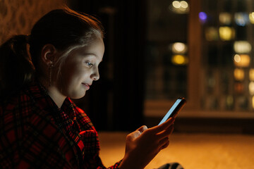 Side view of happy smiling 10yo girl browsing smartphone screen, sitting on bed in cozy dark bedroom with soft bokeh light on blurred background, nighttime cityscape reflecting on window.
