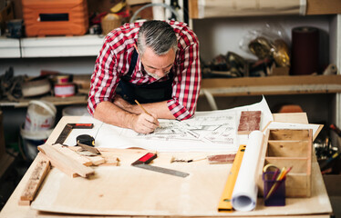 Professional carpenter focused on a blueprint, designing woodwork in a workshop, creating handcrafted furniture with skill and precision in a busy manufacturing environment.