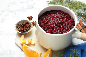 Tasty cranberry sauce in saucepan, spices, orange and fir branches on white marble table, closeup
