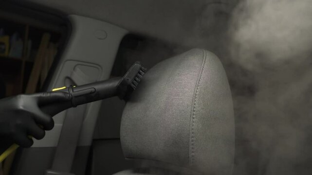 Steam vaping cleaning of car headrest pillow after foam washing with brush 4k video. Worker in car detailing service washing fabric interior of auto using steam generator.