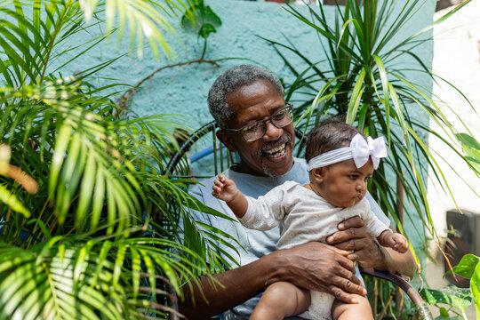 Happy black grandfather with her newborn granddaughter on her lap in a natural environment with plants. Rio de Janeiro, Brazil.