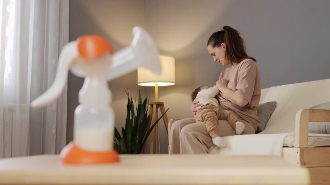 A mother is comfortably breastfeeding her baby in a stylish living room with soft lighting. A breast pump is placed on the table, highlighting a moment of nurturing
