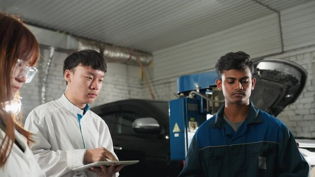 Lab technicians observe engineer demonstrating spanner use on car engine, nodding in agreement in mechanical workshop, industrial workspace features vehicles, tools, and equipment in background