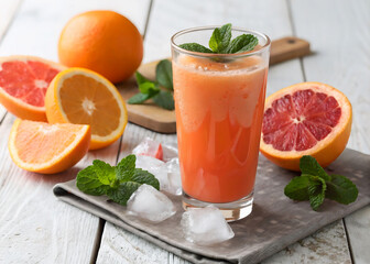 A vibrant display of a freshly squeezed orange juice, garnished with mint leaves, accompanied by halved oranges on a wooden board. Ice cubes are scattered on a slate tray beneath the juice, and the sc