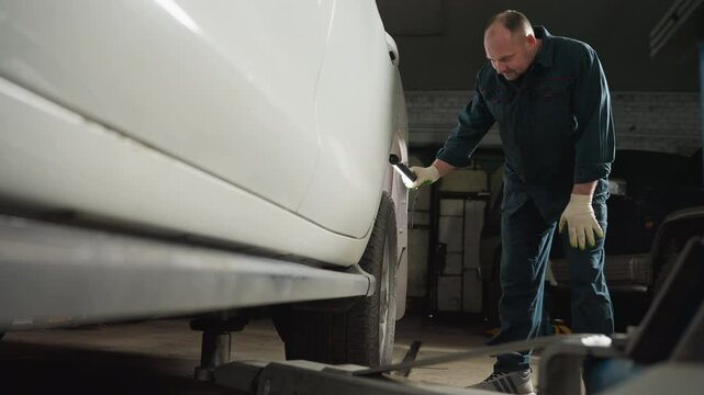 Mechanic in blue uniform uses LED lamp to inspect tire of a white car in garage, bending down with left hand on knee, showing focus and precision
