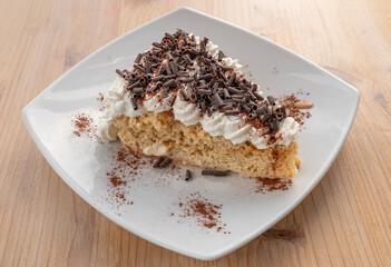 slice of cake with sponge cake base filled with cream and chocolate chips, quick homemade recipe for a snack or party  white plate resting on a wooden table 