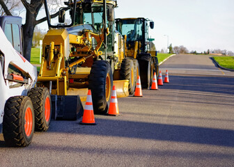 A line of Construction Tractors or Industrial Vehicles parked on a city street surrounded by orange construction cones in a suburban neighborhood.