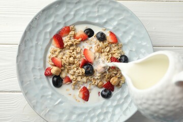 Pouring milk into bowl with oatmeal, berries and hazelnuts at white wooden table, top view