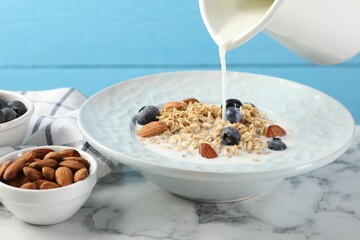 Pouring milk into bowl with oatmeal, blueberries and almonds at white marble table against light blue background, closeup