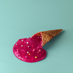 Overturned waffle cone spills bright pink glossy mass with glitter and candy beads on pastel mint background. Surreal food concept