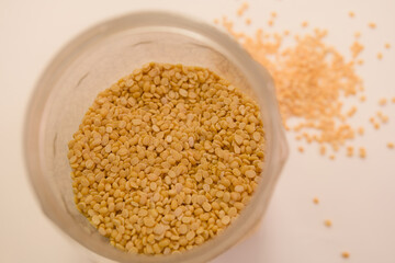 Yellow sesame seeds in glass jar with some scattered on white surface