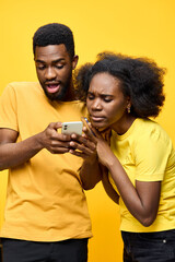 Young couple sharing a moment of surprise together while looking at a smartphone against a bright yellow background, wearing matching yellow outfits