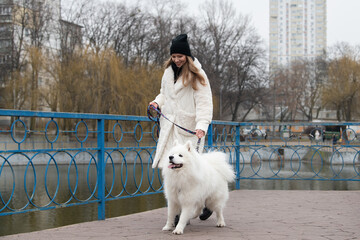 A woman dressed in a warm coat walks her cheerful Samoyed dog on a bridge, surrounded by trees and...