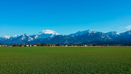 Obraz premium Green meadow stretches towards small village of St. Egyden in Carinthia, Austria. Majestic snow-capped Karawanks mountain range creating dramatic backdrop under pristine clear blue sky. Austrian Alps