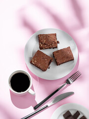 Pieces of chocolate brownie with nuts on a plate with a cup of espresso coffee on a pink background