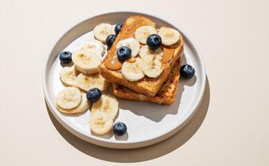 Peanut butter toast on a plate with banana and blueberry on a beige  background with shadow.