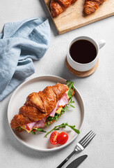Croissant sandwich with ham, cheese, tomato  and arugula on a plate on a light background