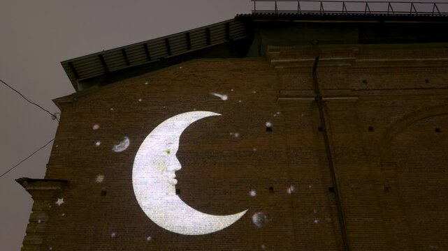 A whimsical moon projection with a face, surrounded by glowing stars, cast onto a historic brick wall in New Holland, Saint Petersburg, creating a magical and artistic evening ambiance
