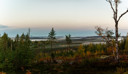 Scenic morning view from hillside overlooking misty valley with autumn forest, distant lakes, and soft pastel sky