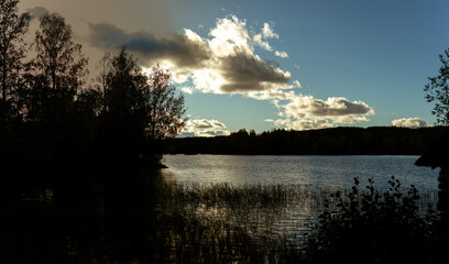 Obraz premium Dramatic Sunset Over Tranquil Lake with Silhouetted Trees and Boathouse in Finnish Wilderness
