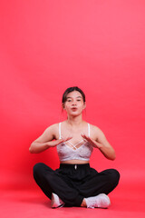 Fototapeta premium A woman is sitting in a cross-legged pose, wearing a sports bra and pants with her hands up, isolated on a red background.