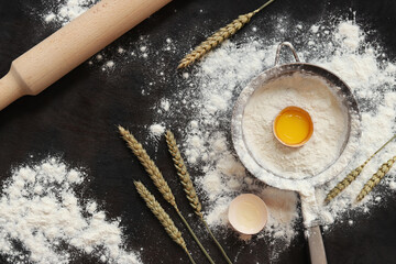 Baking process background, top view. Flour with raw egg, rolling pin for rolling out dough, sieve and ears of wheat. Cooking. Heap of flour on dark table, items for dough preparation. Flat lay