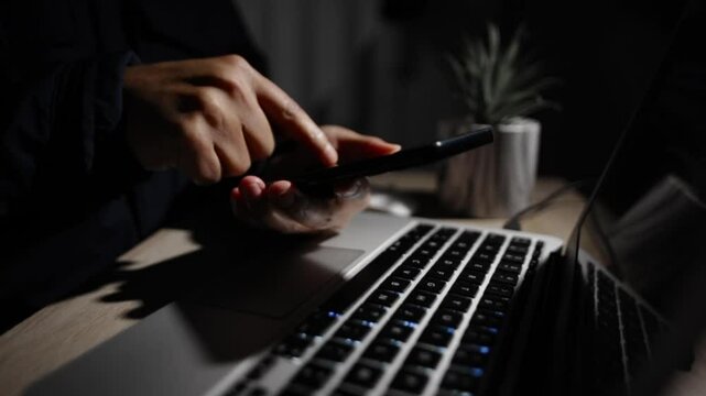 Verifying smartphone code while typing on laptop keyboard, fingertips illuminated by low light digital workspace screen