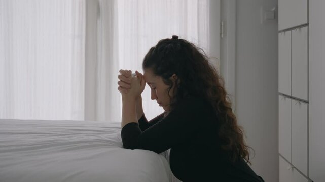 Young woman praying by the bed kneeling on floor with hands clasped together. Religious female holds hands and with eyes closed prays or worships indoors in apartment or house during daytime