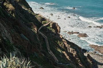 Wooden staircase descends a dramatic cliffside towards a vibrant ocean. Waves crash against the rocky shore creating a breathtaking coastal scene. Perfect for travel, adventure, and nature themes.