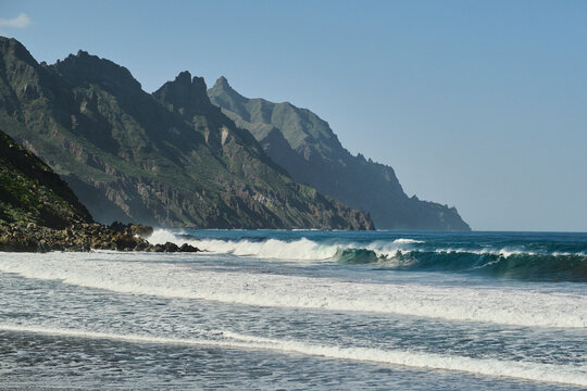 Dramatic coastal landscape with powerful ocean waves crashing on a rocky shore, under a clear blue sky. Rugged mountains rise majestically in the background, creating a stunning natural scene.
