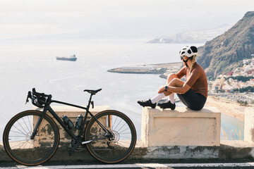 Female cyclist takes a break on a scenic coastal overlook, enjoying the view with her road bike. Stunning ocean backdrop and sunny weather.