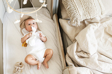 Cute little baby lying in crib with sweet toy and boho mobile. Bedtime. Adorable infant portrait in...