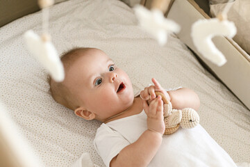 Cute little baby lying in crib with sweet toy and boho mobile. Bedtime. Adorable infant portrait in...