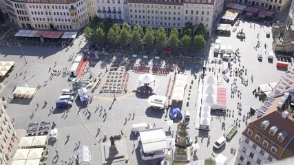 The bustling market square is filled with colorful stalls, tall trees, and people enjoying the lively atmosphere of a European city