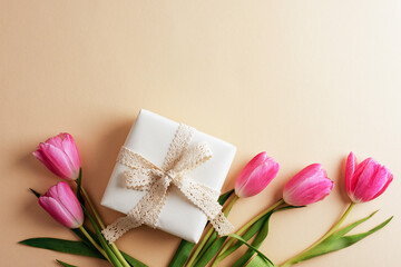 Wrapped gift box with a lace ribbon surrounded by pink tulips on a beige background. Womens day concept. Top view, flat lay
