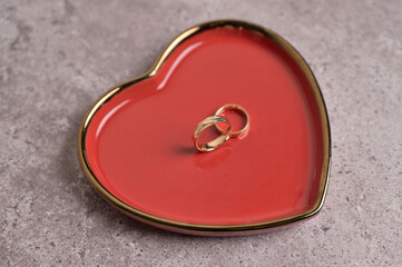 Couple of wedding golden rings on red heart shape ceramic plate with a gold rim. Gray concrete...