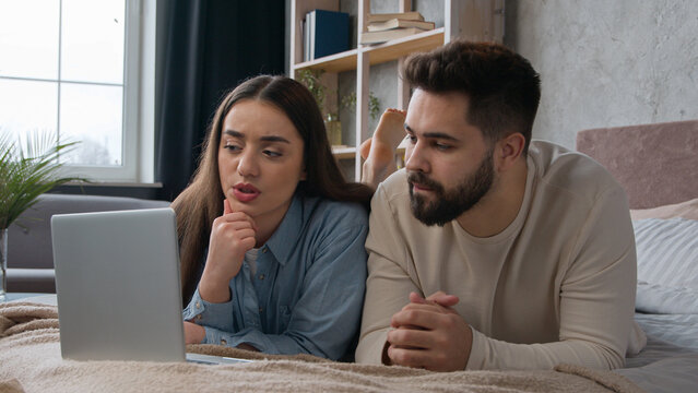 Caucasian couple homeowners family thoughtful husband and wife shopping online on bed choose apartment on laptop application doubtful man and woman talk thinking planning internet purchase in bedroom