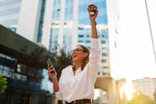 Businesswoman celebrating success with coffee and smartphone in city