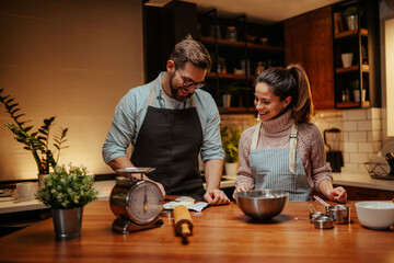 Happy couple wearing aprons preparing food in kitchen using scale and reading recipe