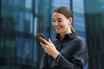 Young woman smiling while using smartphone outside a modern glass building in daylight